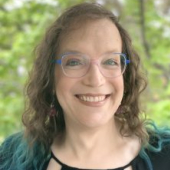 Stephanie Burt, a Jewish woman with shoulder-length, curly hair that is medium brown with teal tips, in a head-and-shoulders photo. She is wearing dangle earrings, trans-flag glasses that fade from blue to white to pink, and a black top with cut-outs. She is smiling at the camera. Headshot of Stephanie Burt, who is wearing glasses and smiling.