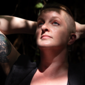 Sarah Gailey, a white person with closely shaved hair on the sides and longer hair on top, gazes upward with their hands resting behind their head. They are partly lit by sunlight, showing a tattoo on their upper arm, against a dark leafy background. © Sarah Gailey