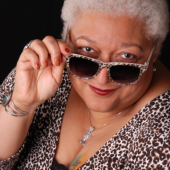 Jewelle Gomez, a woman of Cabo Verdean, Wampanoag, and Ioway heritage, with short, white, curly hair. She is wearing cheetah-print sunglasses, a silver pendant, a silver gemstone bangle on her right wrist, a silver ring on her right ring finger, a black tank top, and a long-sleeved cheetah-print top. The black, green, and gold tattoo on her left breast is partly visible. Her right hand is holding her glasses partway down her nose, and she is looking saucily over them at the camera. 
A headshort of Jewelle Gomez, who is holding her sunglasses partway down her nose to look over them at the camera.
© Irene Young 2013