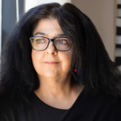 Layla Azmi Goushey, a Palestinian-American woman with long, black hair parted in the center, in a head-and-shoulders photo. She is wearing black glasses, red-and-black dangle earrings, and a black top with an embroidery-style red design across the front. She is looking off in the distance with a slight smile. 
A headshot of Layla Azmi Goushey, who is looking off into the distance with a slight smile.