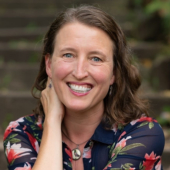 Kelly Barnhill, seated, faces the camera and smiles, leaning her elbows on a knee.